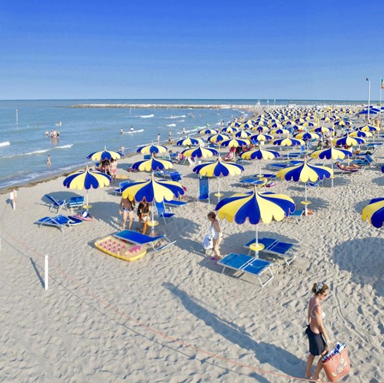 Spiaggia affollata con ombrelloni colorati, mare calmo e vegetazione rigogliosa sullo sfondo.