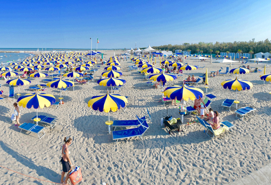 Spiaggia affollata con ombrelloni gialli e blu, mare calmo e cielo sereno.